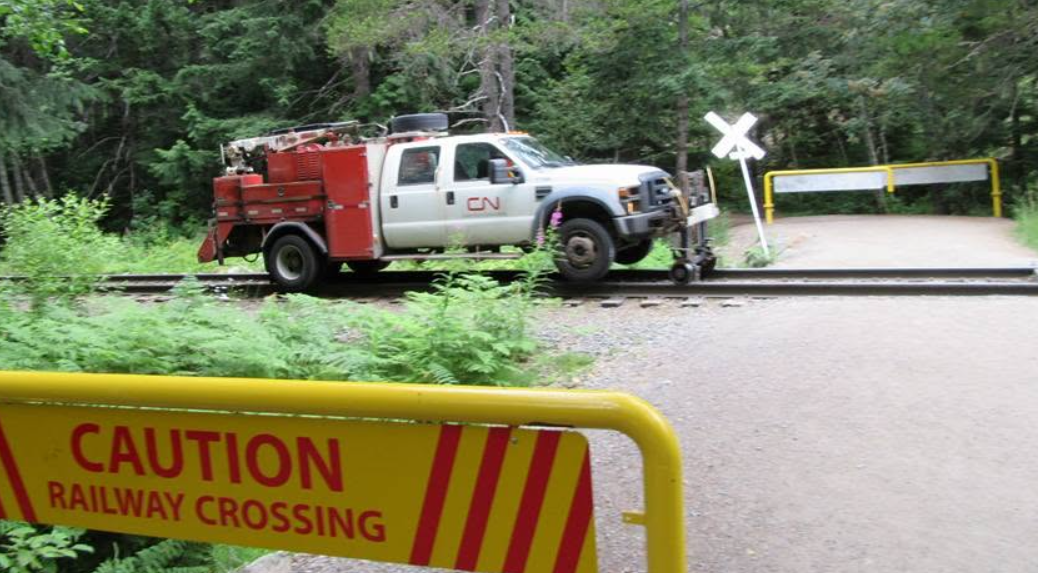 A CN hi-rail maintenance truck straddling a railway crossing, with a yellow CAUTION RAILWAY CROSSING gate in the foreground